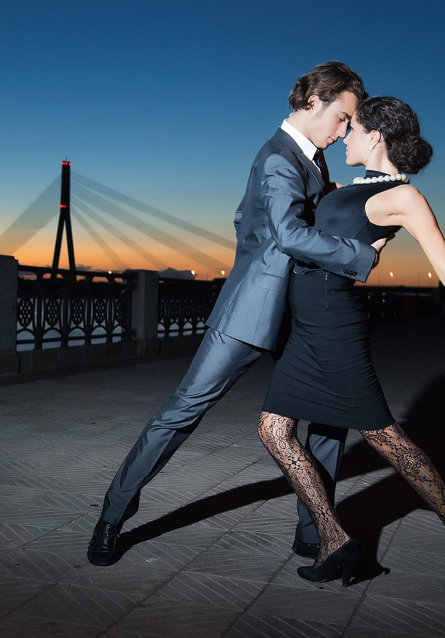 A couple in a close tango embrace on a riverside promenade at sunset, a bridge behind them