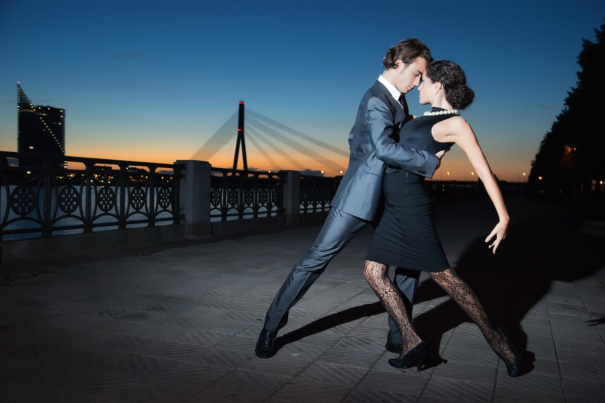 A couple in a close tango embrace on a riverside promenade at sunset, a bridge behind them