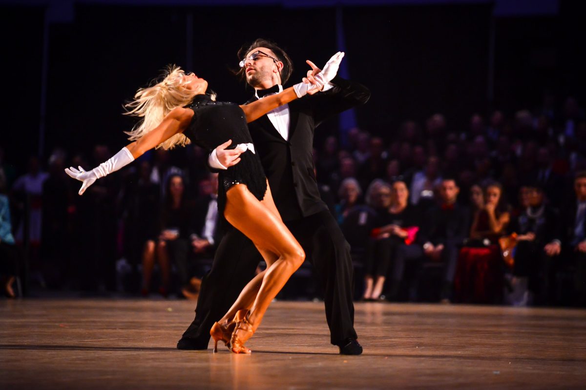 A ballroom couple in a sweeping pose on a competition floor, the woman in long white gloves