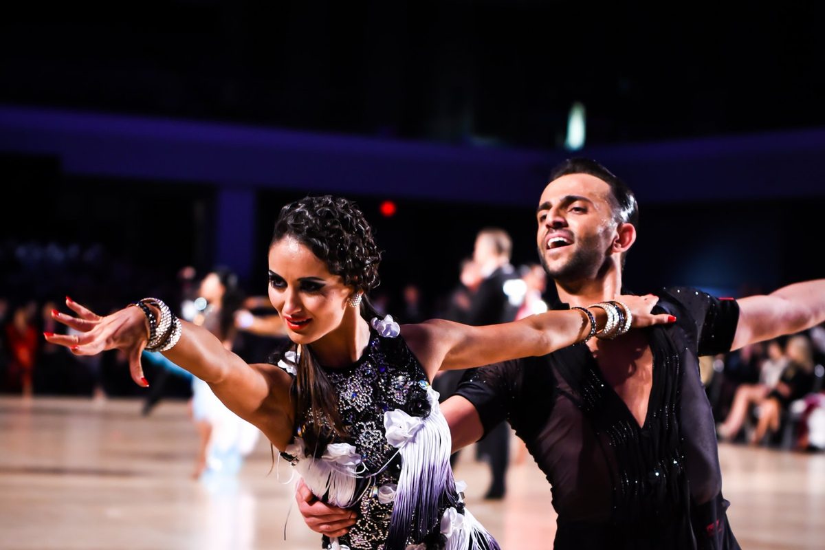 A Latin couple on a competition floor, arms extended, the woman in a black and white fringed dress