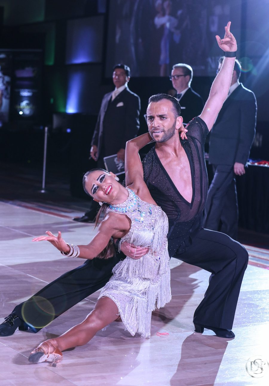 Vard Margaryan in a deep Latin lunge with his partner in a white fringed dress on a competition floor, judges looking on