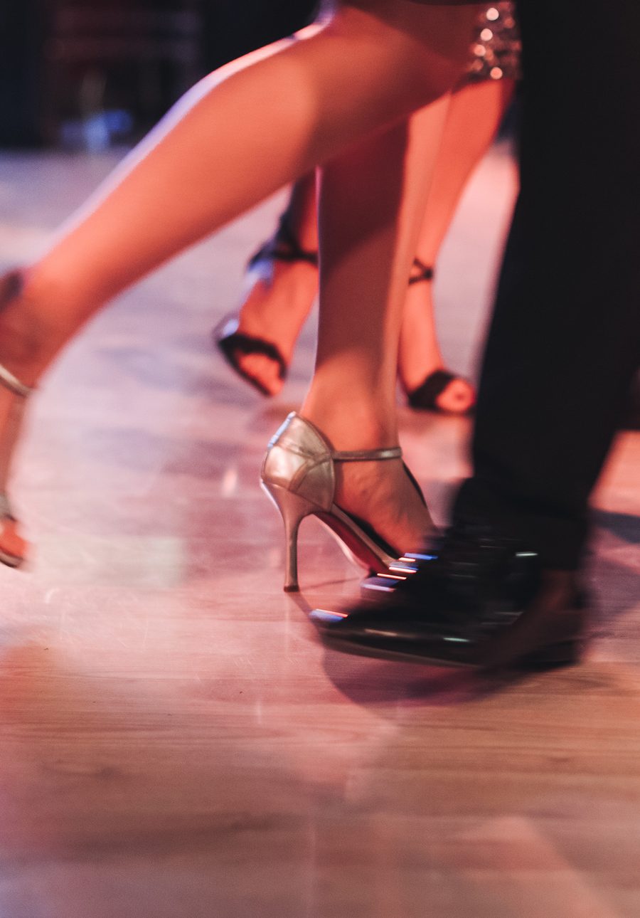Close view of a couple’s feet mid-step on a party dance floor, her silver heels and his patent shoes