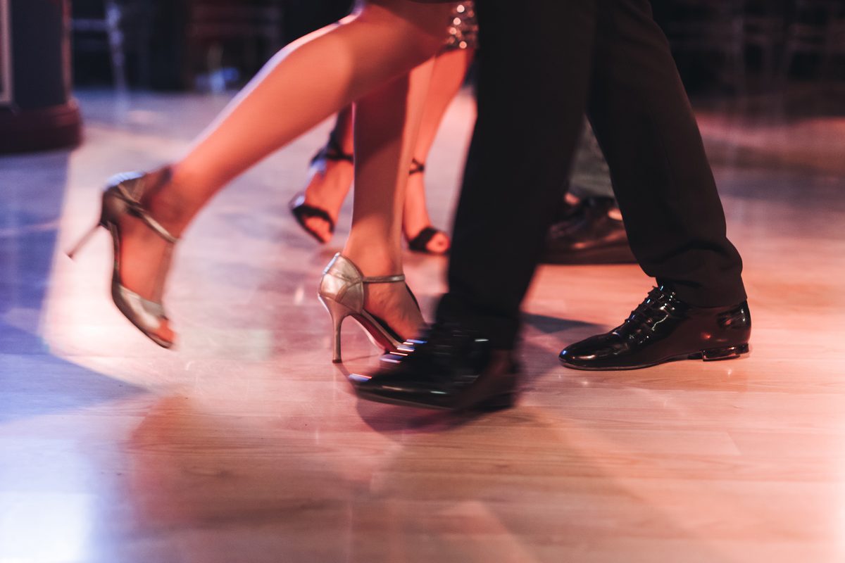Close view of a couple’s feet mid-step on a party dance floor, her silver heels and his patent shoes