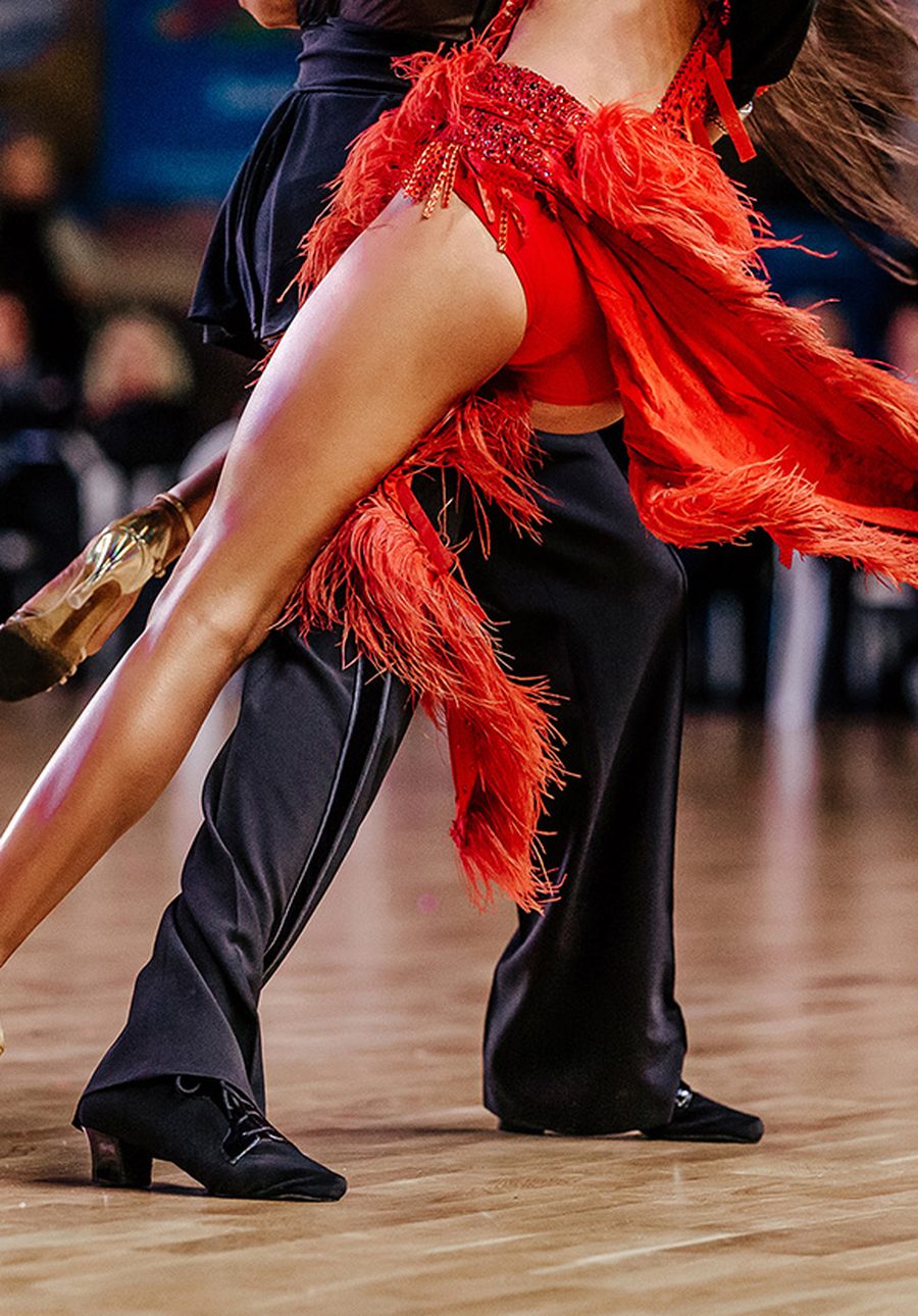 A Latin dancer in a red fringed dress kicking out beside her partner on a competition floor