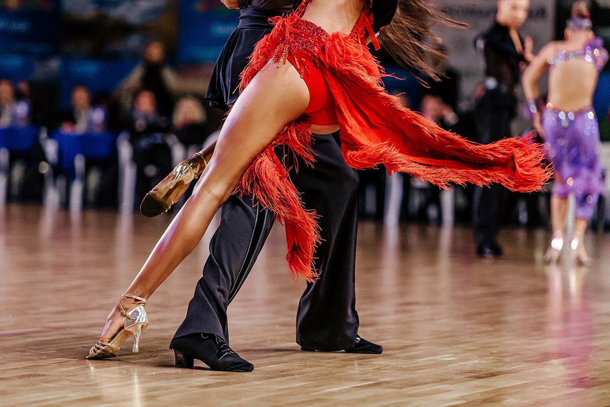 A Latin dancer in a red fringed dress kicking out beside her partner on a competition floor