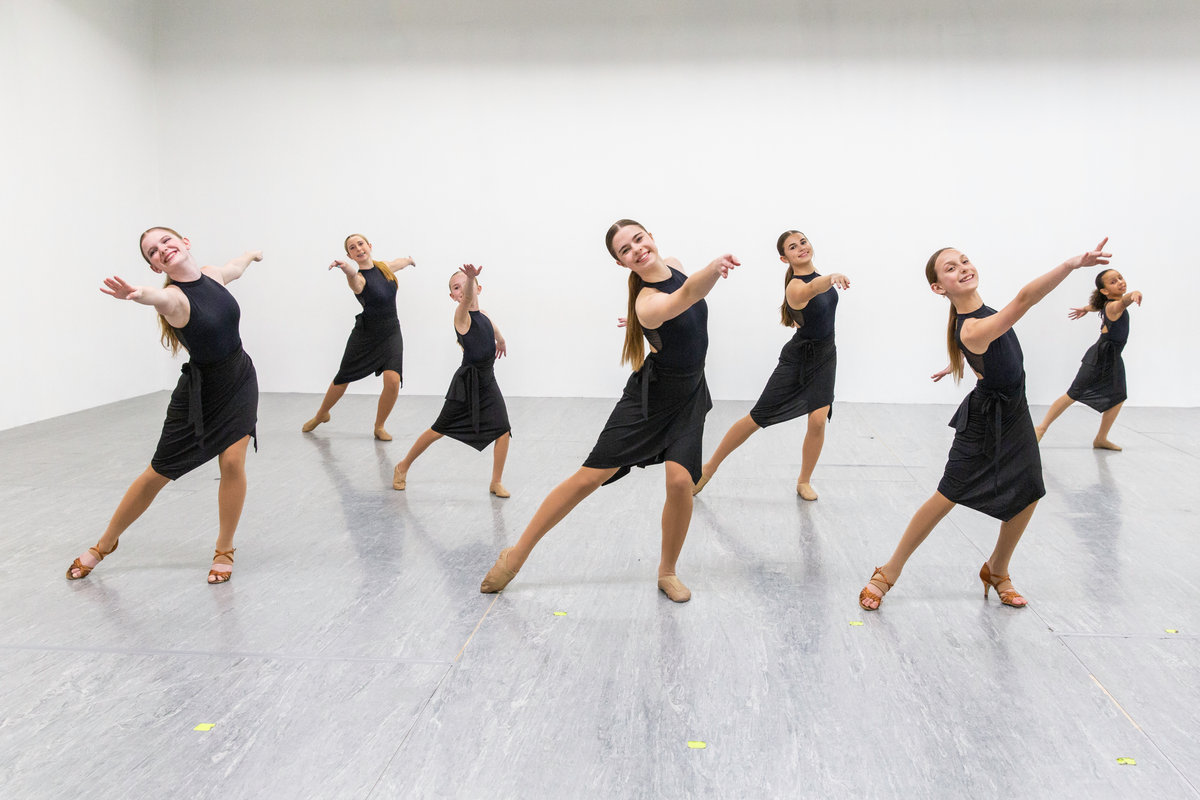 A line of young dancers in black dresses striking the same pose across the studio floor