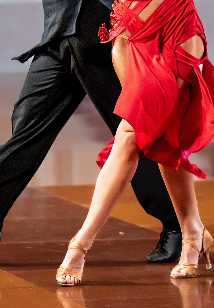 A couple’s legs mid-step on a polished floor, the woman in a red Latin dress and tan dance shoes