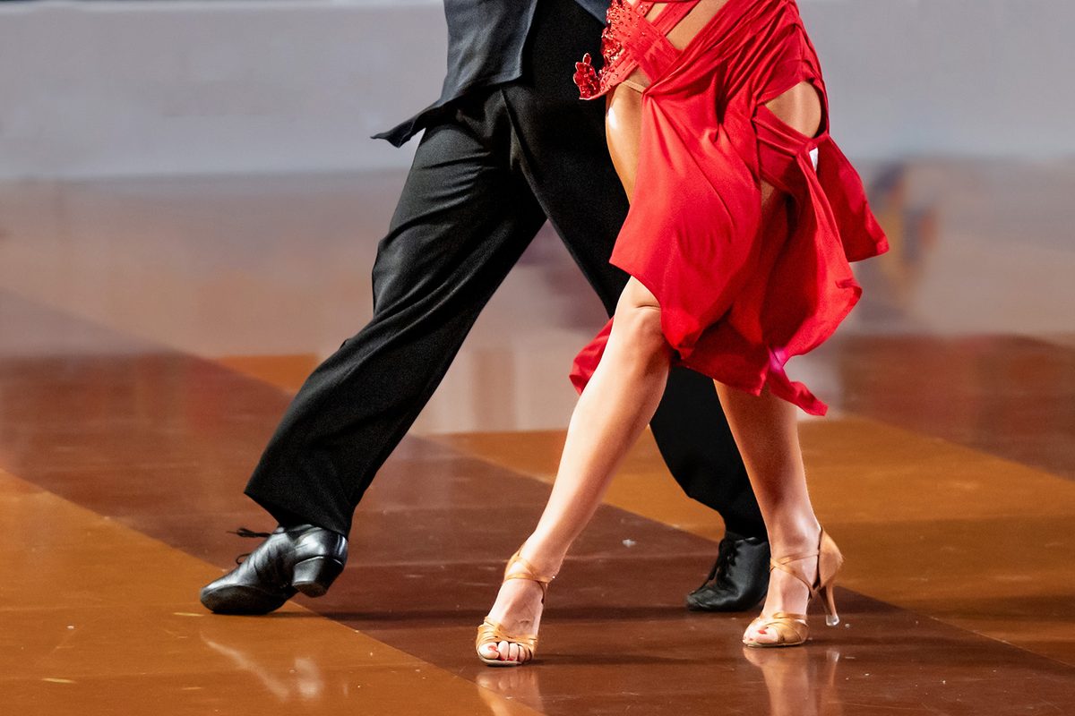 A couple’s legs mid-step on a polished floor, the woman in a red Latin dress and tan dance shoes