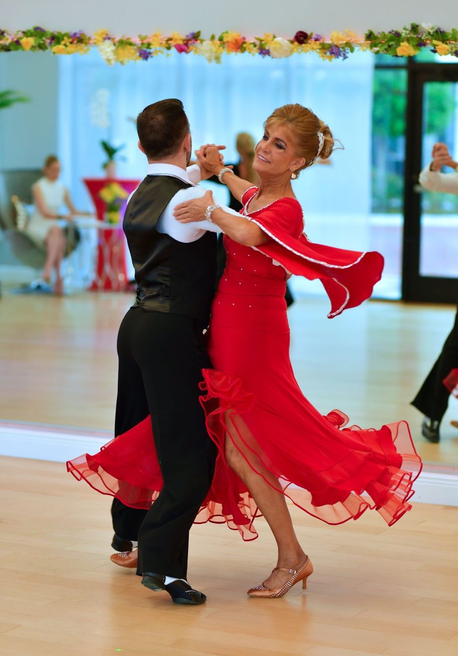 An instructor and a student in a red gown with flared sleeves travelling across the VM Ballroom floor