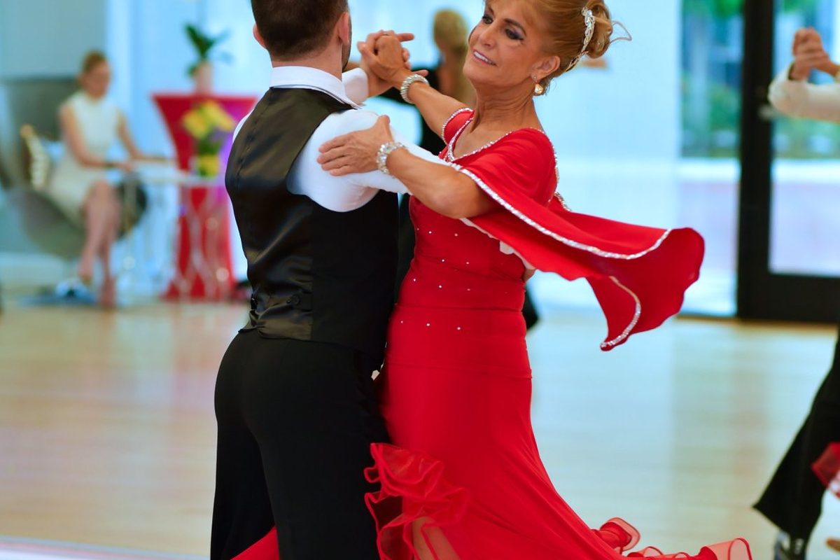 An instructor and a student in a red gown with flared sleeves travelling across the VM Ballroom floor