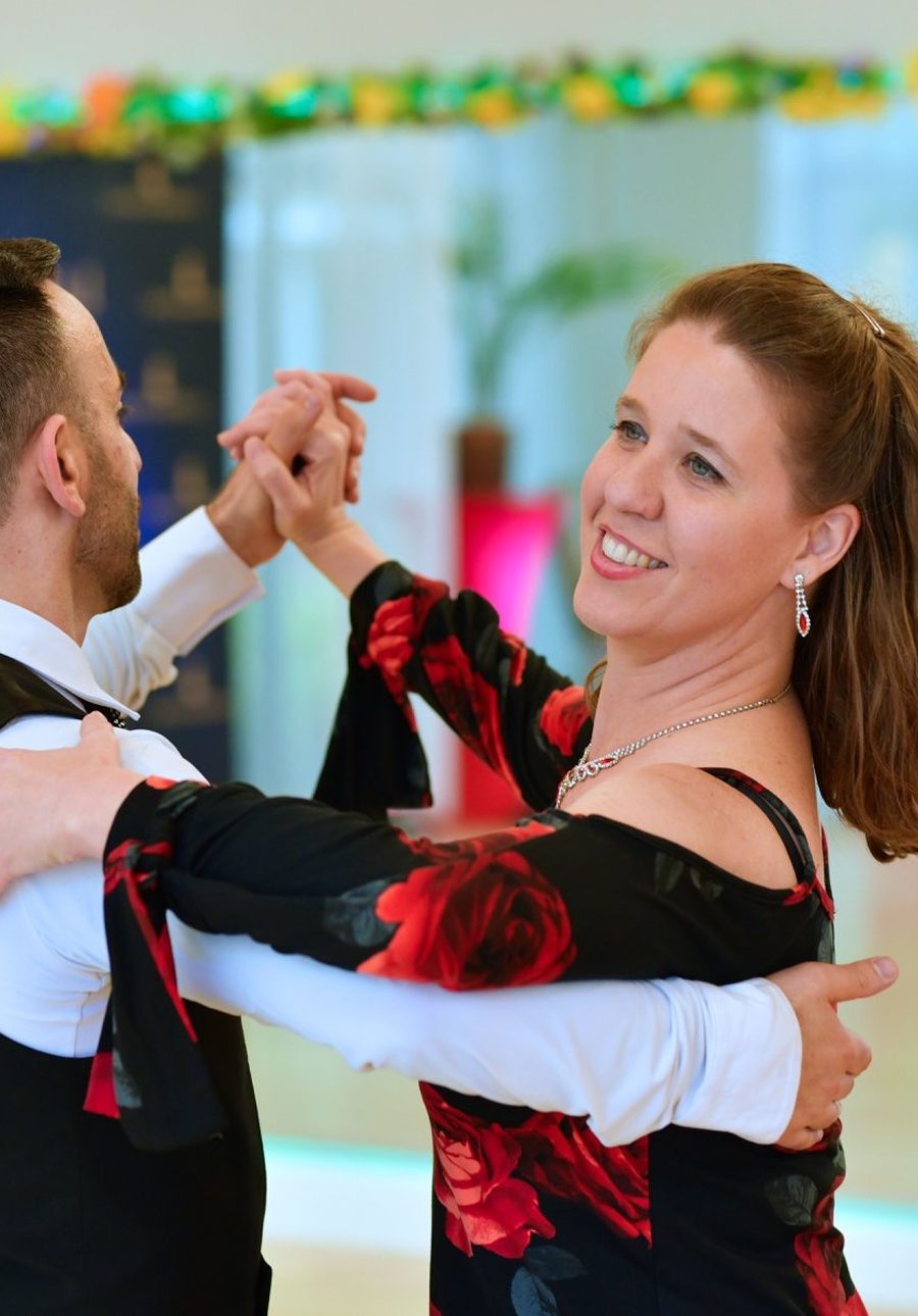 An instructor and a smiling student in a black gown with red roses dancing in closed hold at VM Ballroom