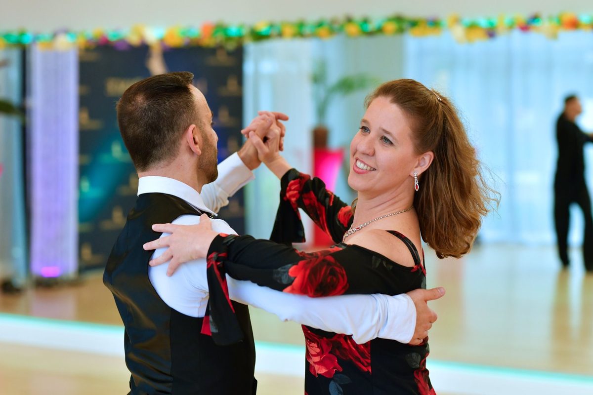 An instructor and a smiling student in a black gown with red roses dancing in closed hold at VM Ballroom
