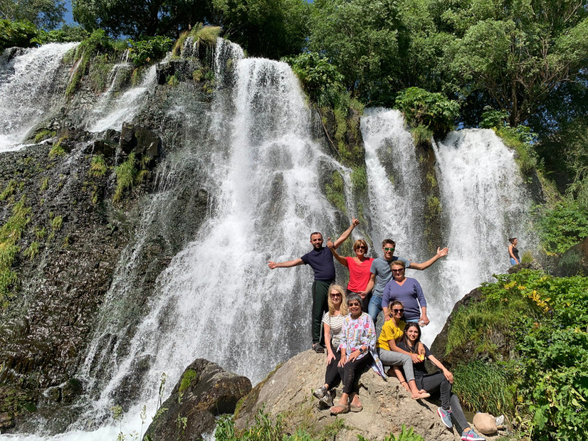The group beside a wide waterfall falling over a mossy cliff, arms raised