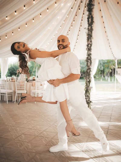 A couple in white dancing under a draped canopy strung with lights, the follower leaning back on her partner's arm