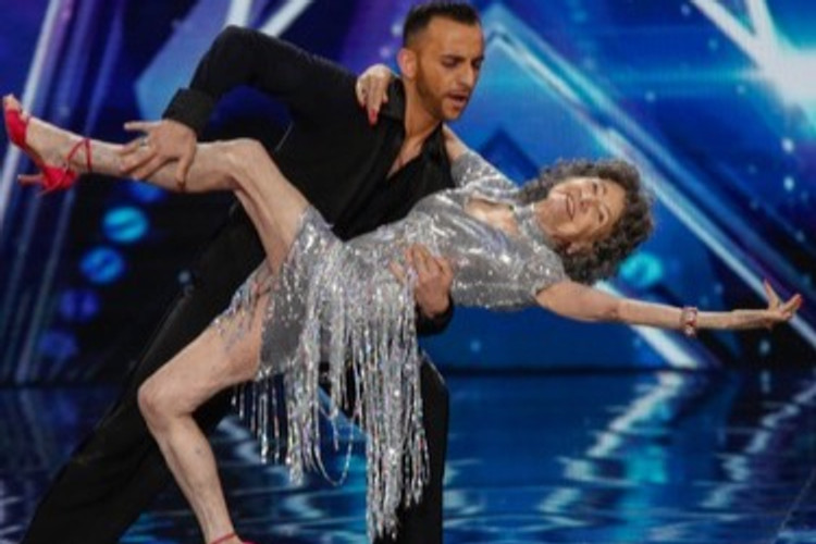 A dancer in a fringed silver dress held horizontally in a deep lunge by her partner under stage lights