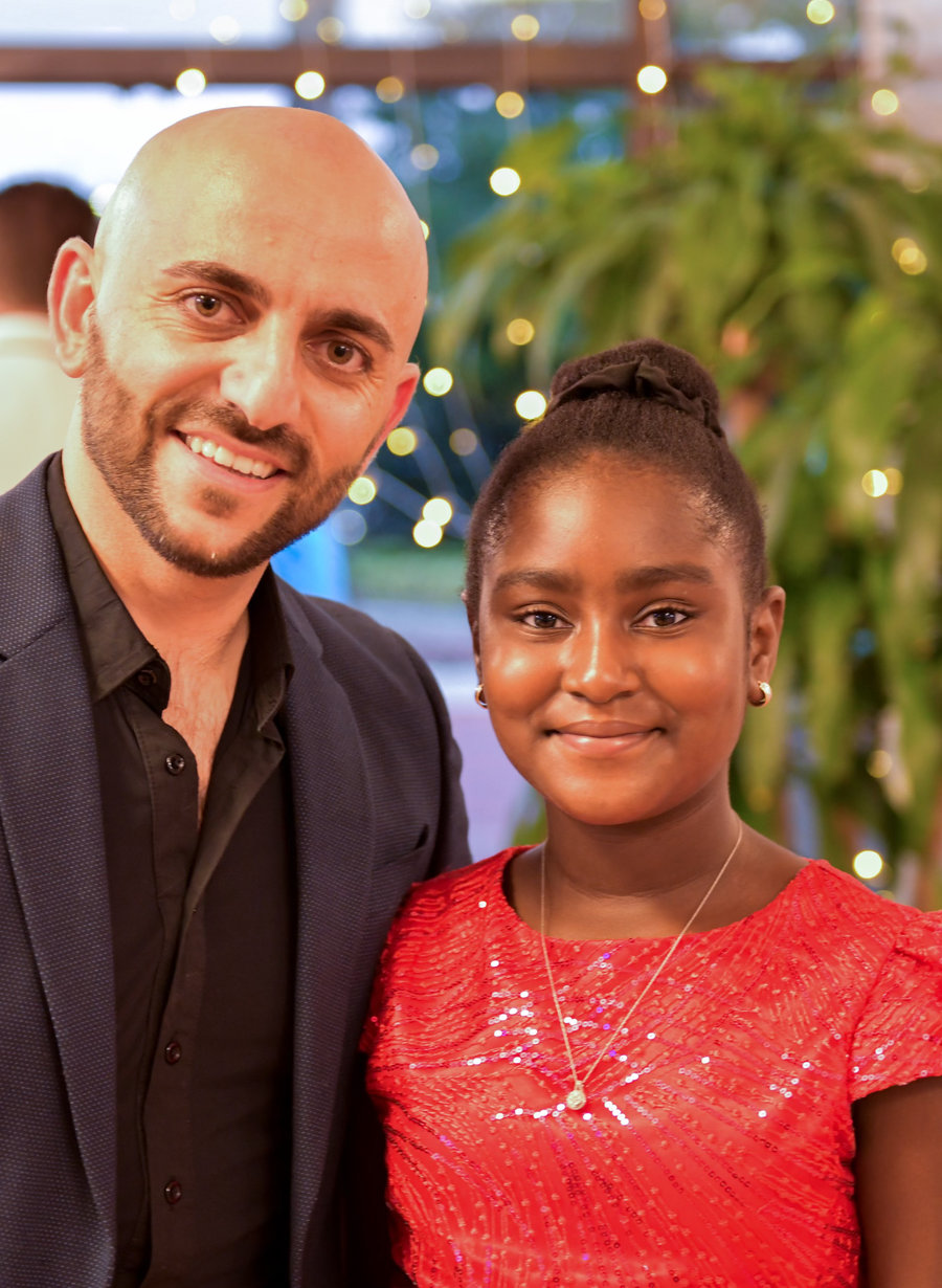 An instructor and a young student in a red sequinned dress smiling together at a party in the VM Ballroom Dance Studios ballroom, under strings of lights