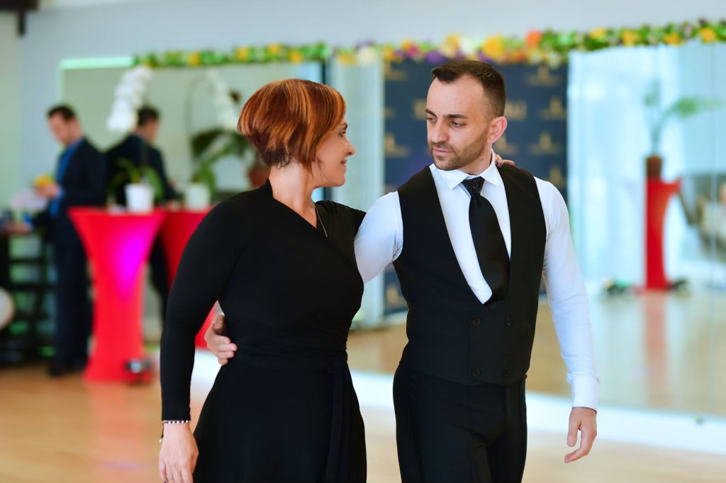 A woman and a man standing together in dance hold on the floor at VM Ballroom Dance Studios, with the ballroom's tall windows behind them