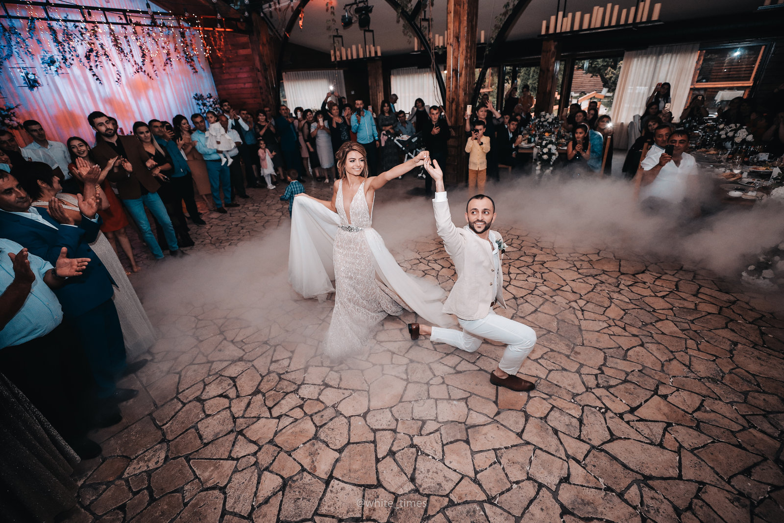 A bride in a long white gown and a groom in a white suit dancing their first dance in front of their guests, lit from behind through a cloud of dry ice