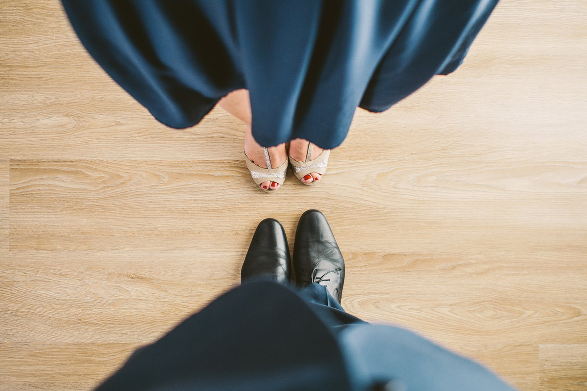 Looking down at a couple’s dance shoes on a wooden floor, the woman in a long navy gown