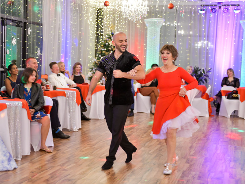 An instructor and a student in a red dress dancing apart in open position at a studio party, a lit Christmas tree and guests at tables behind them