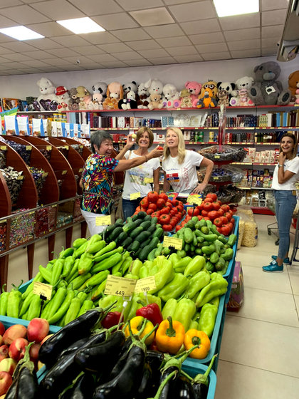 Travellers in a food shop behind crates of peppers, cucumbers and tomatoes, with shelves of sweets and dried fruit behind them