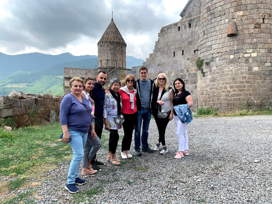 The party on the grass below a hilltop monastery with a conical-domed church and stone walls, mountains behind