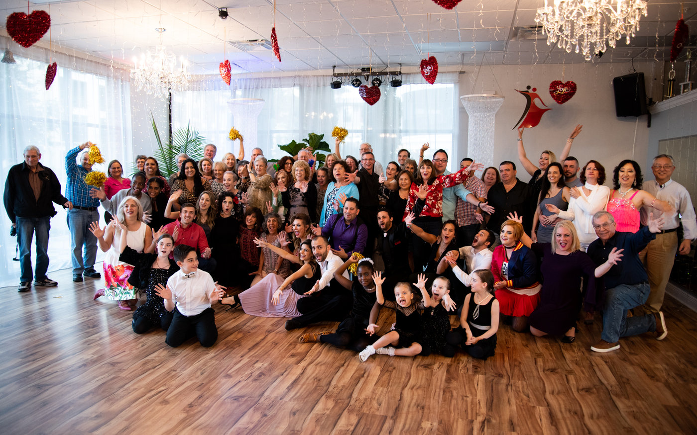 The whole studio gathered on the floor at VM Ballroom Dance Studios — students, families and children together, hands in the air, under hearts and chandeliers