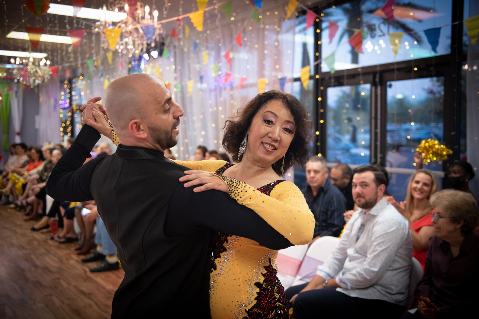 An instructor dancing with a student in a gold dress while guests watch from the tables at a VM Ballroom Dance Studios party