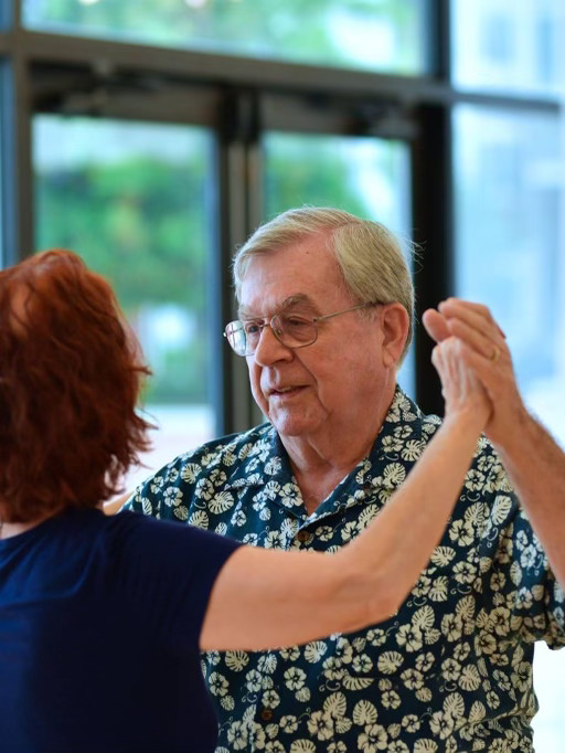 An older gentleman in a patterned shirt dancing in hold with his partner in a bright studio