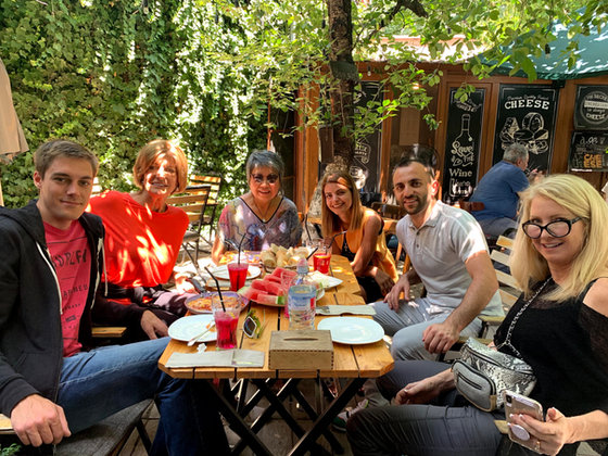 Six of the group around a cafe table under a vine, with watermelon and cold drinks