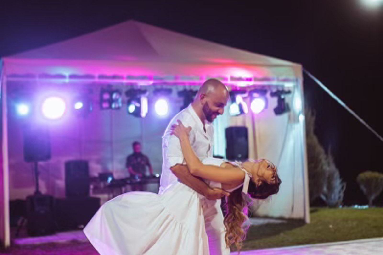A couple dancing together under a lit marquee at an outdoor evening event, with a band playing behind them