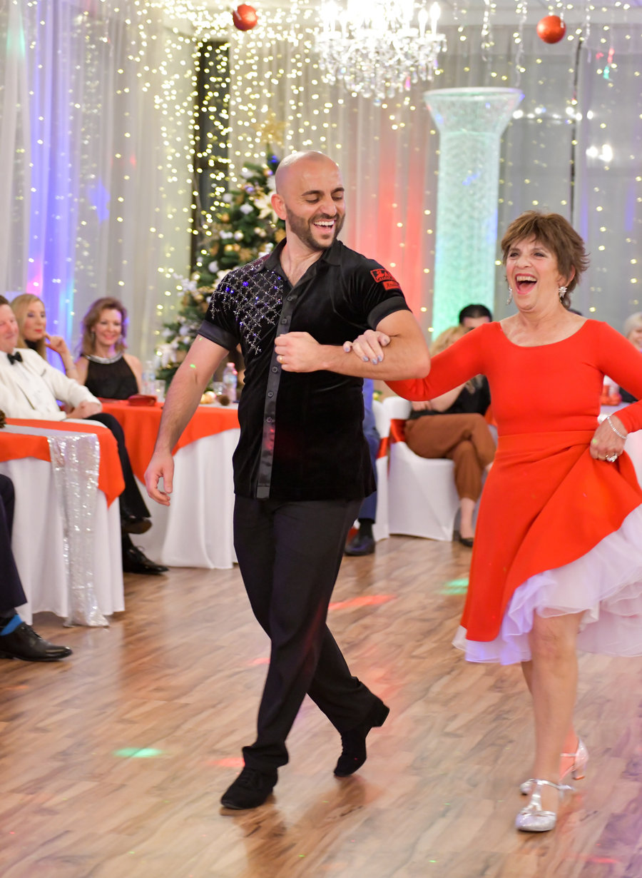 A student in a red dress performing with her instructor at a VM Ballroom Dance Studios showcase while guests watch from tables strung with lights