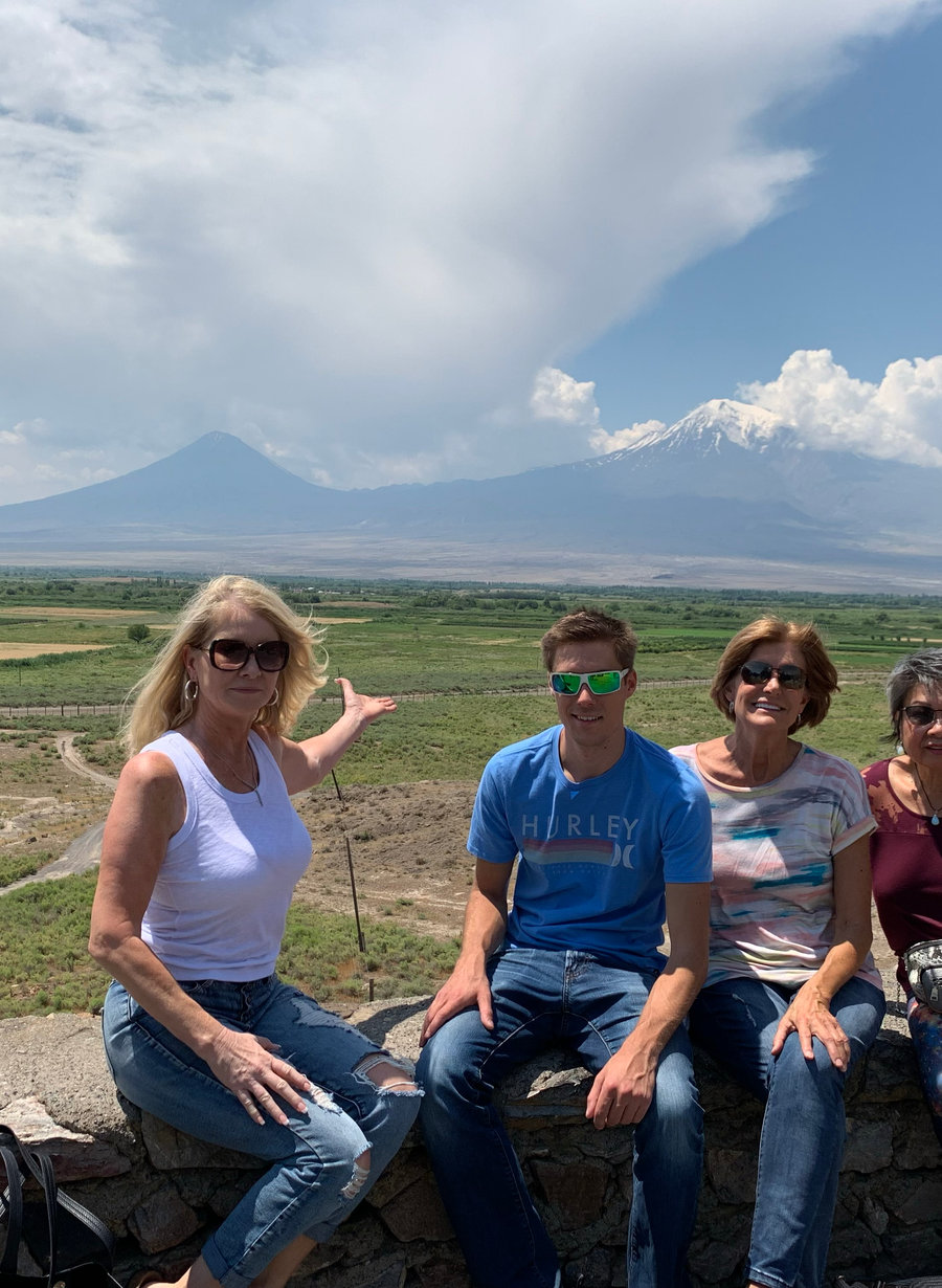 Four of the VM Ballroom Dance Studios travelling party sitting on a stone wall above a green plain in Armenia, a snow-capped mountain on the horizon behind them