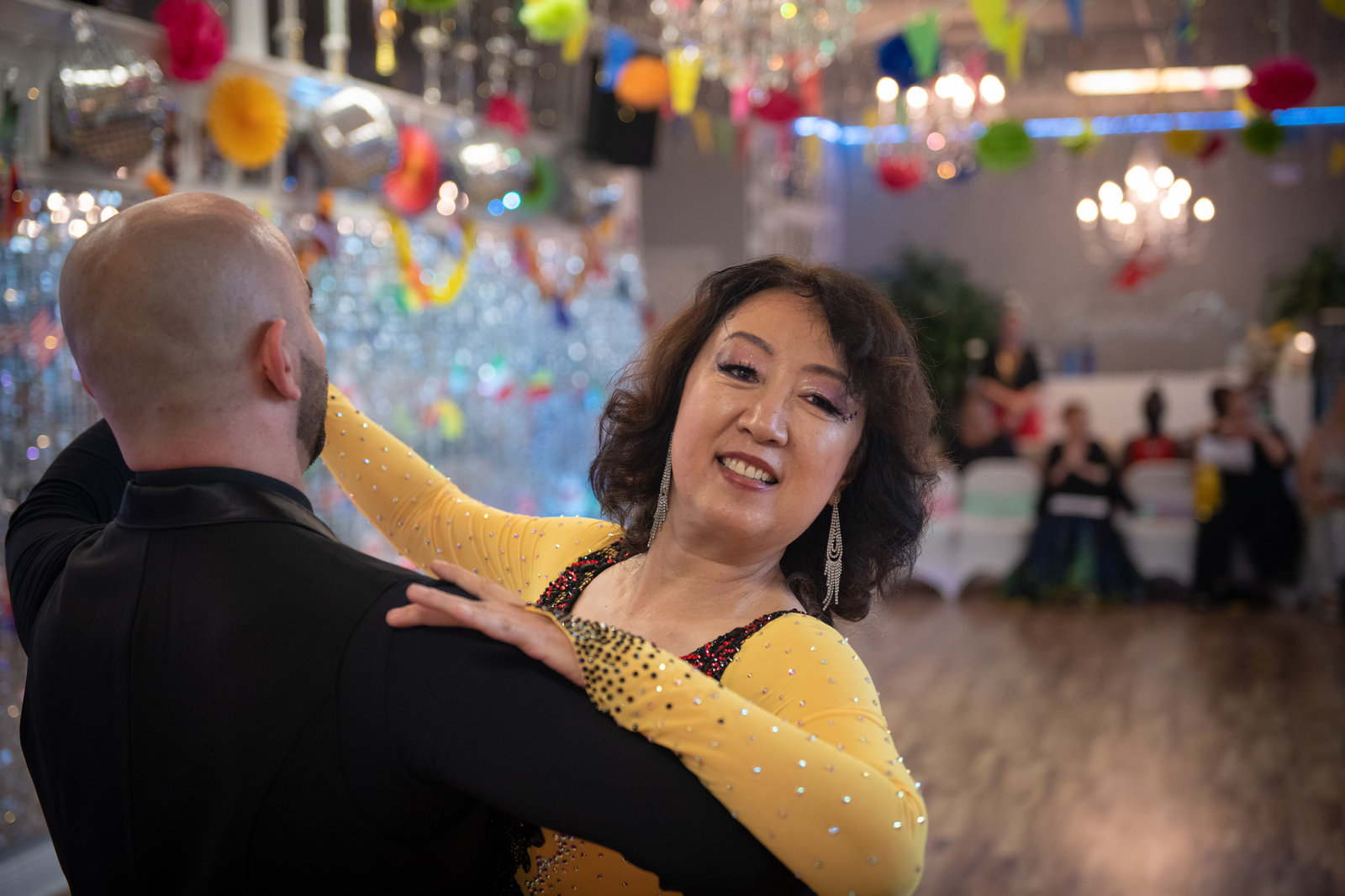 A couple dancing at a party on the VM Ballroom Dance Studios floor, with paper decorations and bunting strung across the room beneath two chandeliers