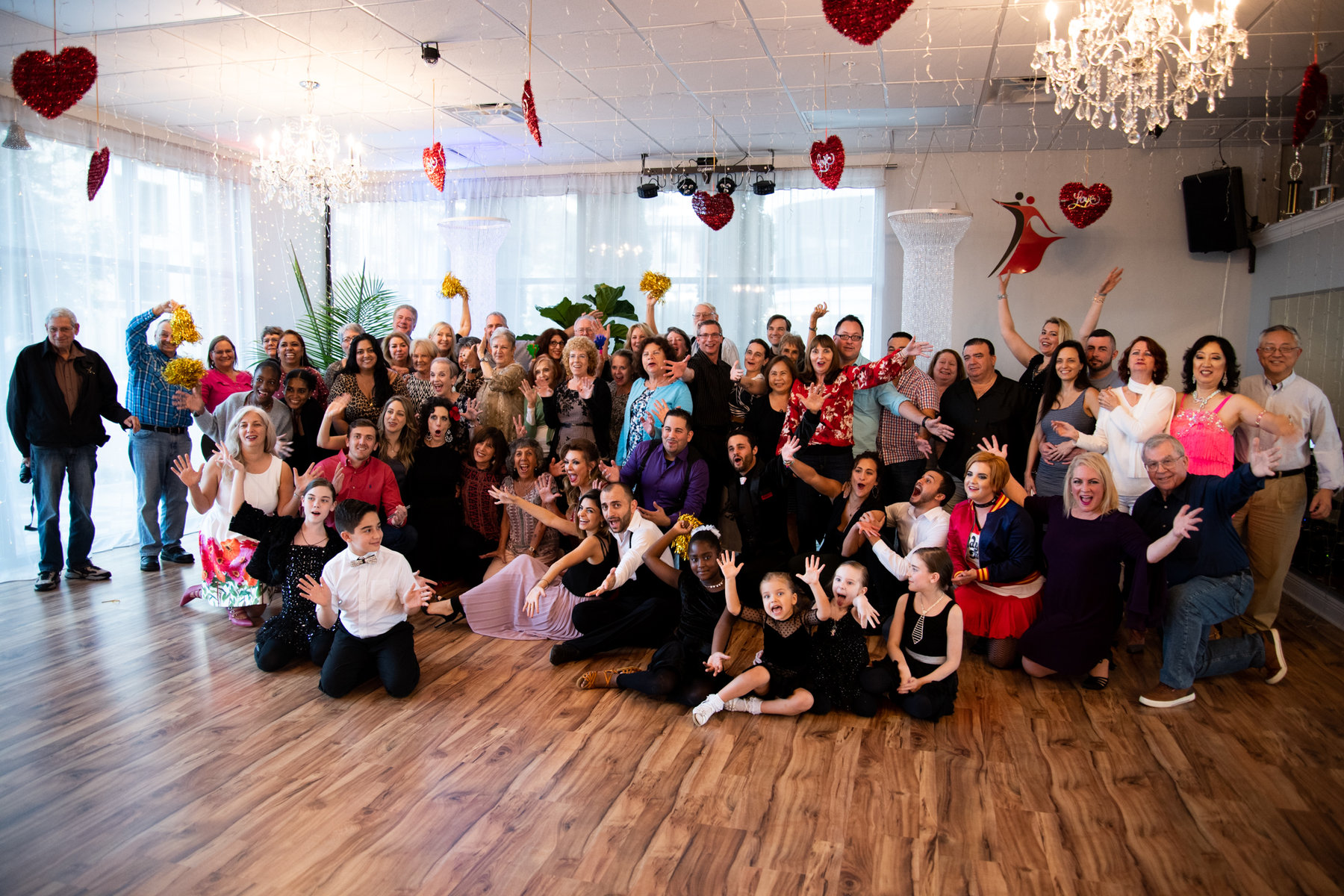 A large group of VM Ballroom Dance Studios students, children, families and instructors gathered together on the studio floor, cheering with their hands in the air beneath the chandeliers and red heart decorations of a studio party