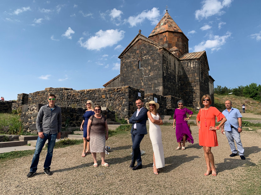 Nine of the travelling party in front of a dark stone church with a conical dome and old stone walls in Armenia