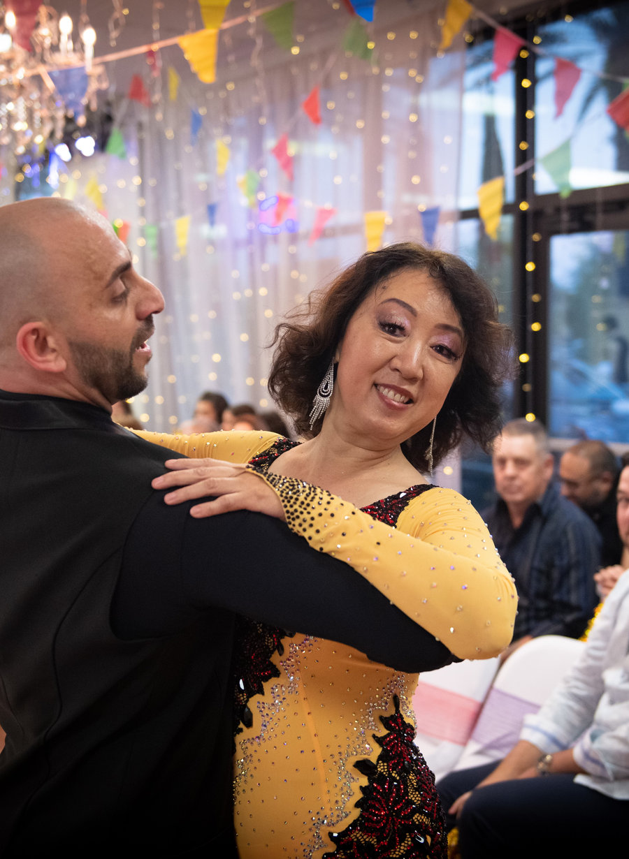 An instructor dancing with a student in a gold dress while guests watch from the tables at a VM Ballroom Dance Studios party